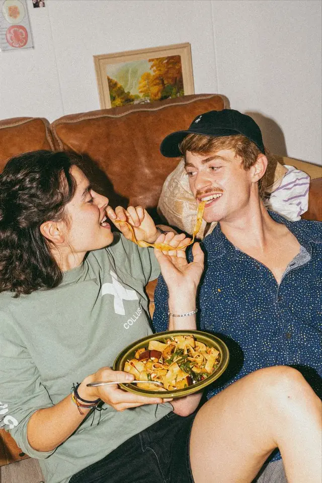 Couple playfully sharing pappardelle on a brown leather sofa