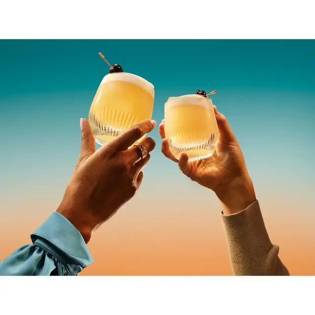 Two frothy cocktails in faceted lowball glasses against teal-to-peach gradient