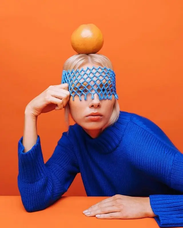 Blue knit sweater, orange balanced on head — high‑contrast studio portrait