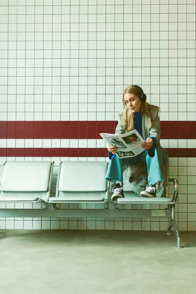 Woman reading magazine on metal waiting-room bench against tiled wall