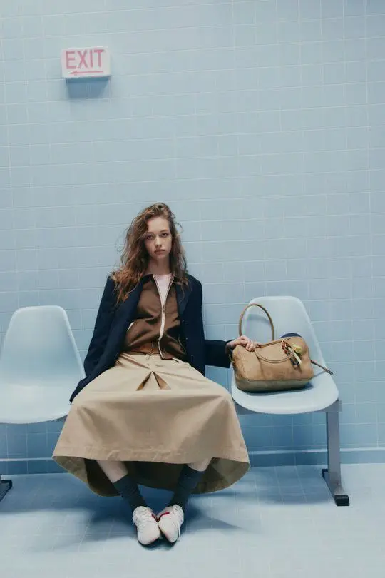 Model seated in pastel blue tiled waiting room with tan leather handbag