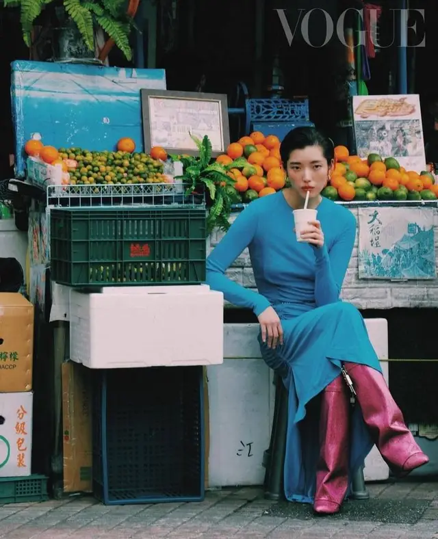 Vogue editorial: model in teal dress and metallic pink boots at a street fruit stall