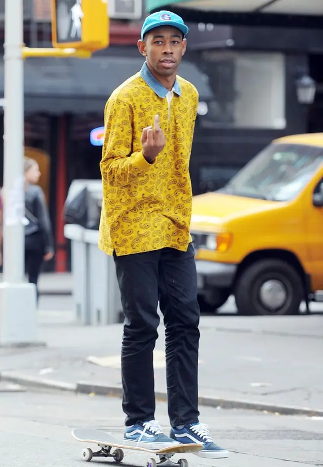 Skateboarder in mustard paisley shirt and teal cap on city street
