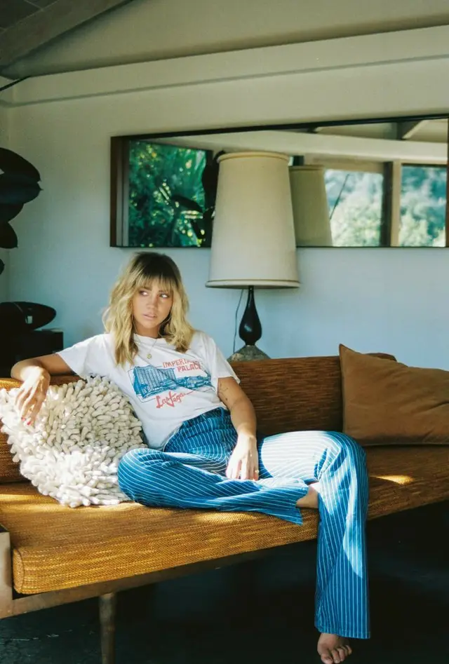 Relaxed mid-century living room — woman on mustard woven sofa in graphic tee and blue pin‑