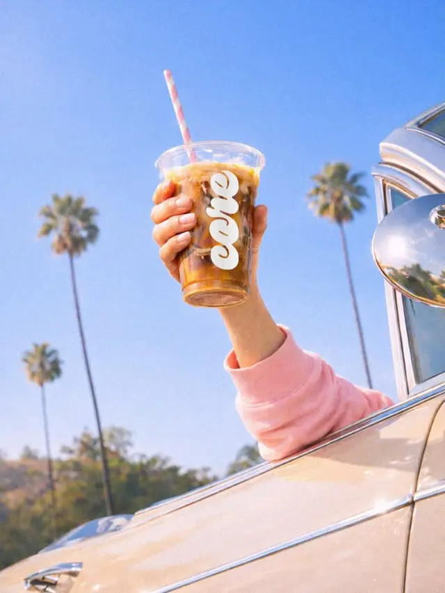 Hand holding iced coffee from car window with palm trees and blue sky