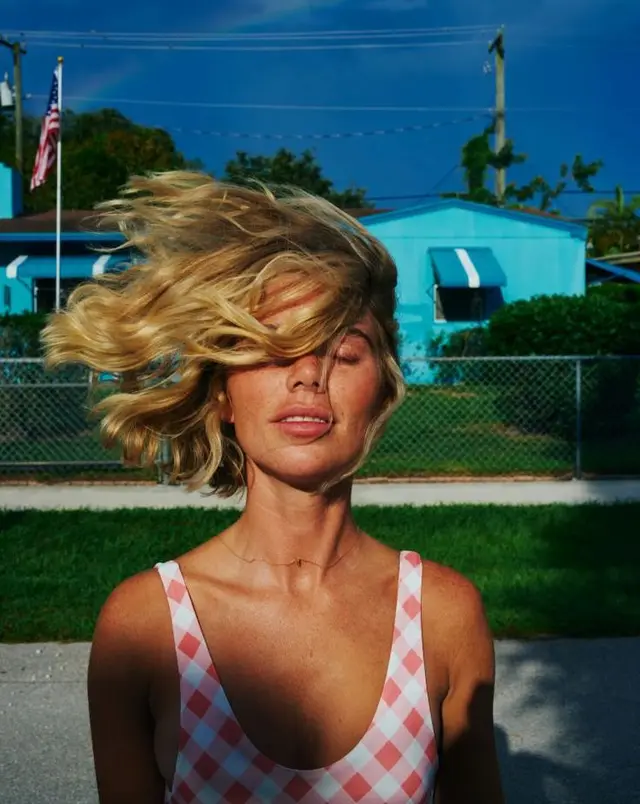 Wind‑blown portrait in pink gingham swimsuit before turquoise bungalow