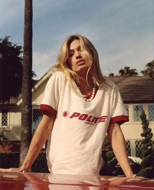 Blonde model in 'POLITE' ringer T-shirt leaning on red car with suburban house and palm