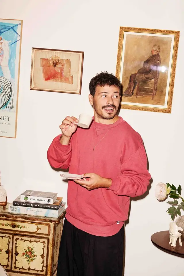 Man in coral sweatshirt holding espresso cup in eclectic interior