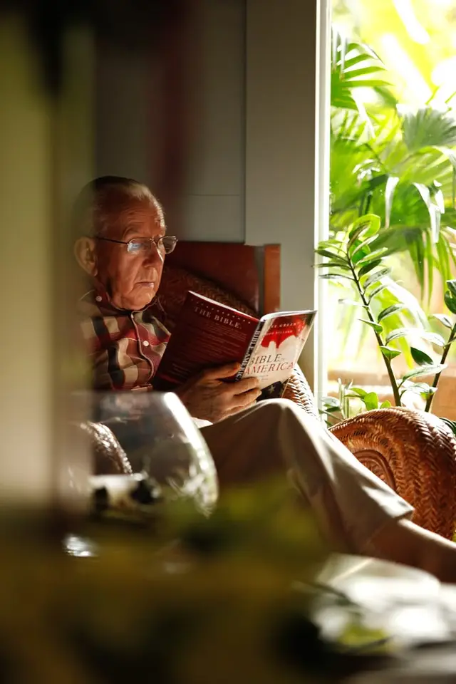 Elderly man reading a hardcover Bible in wicker chair by sunlit window