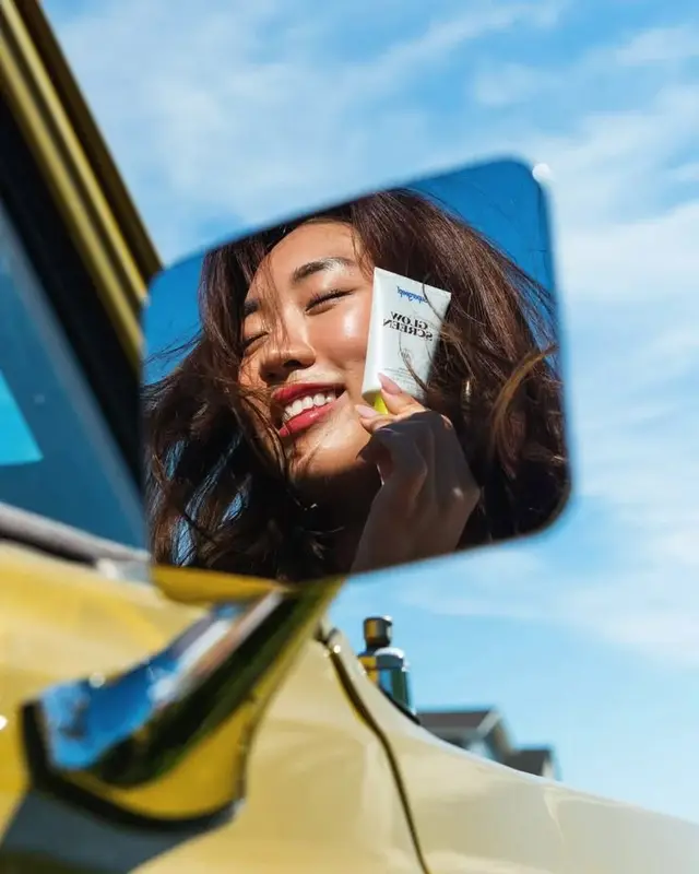 Reflection: woman holding white skincare tube in yellow car mirror