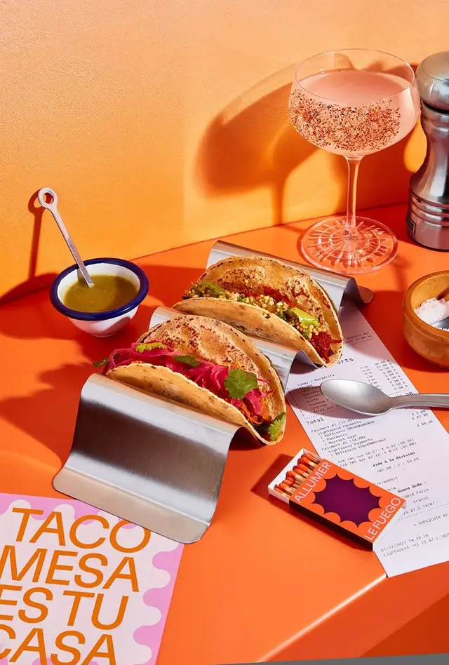 Three tacos on stainless-steel rack with pink cocktail on orange tabletop