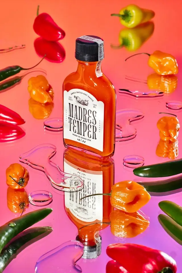 Madres Temper hot sauce bottle on glossy pink mirror with assorted peppers