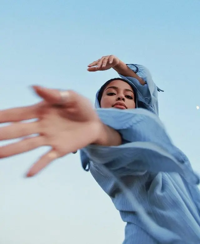 Low-angle portrait of woman in light-blue shirt and headscarf reaching toward camera