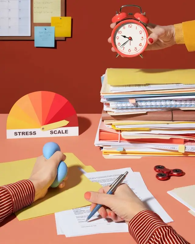 Desk scene with red alarm clock, stress scale and stacked paperwork