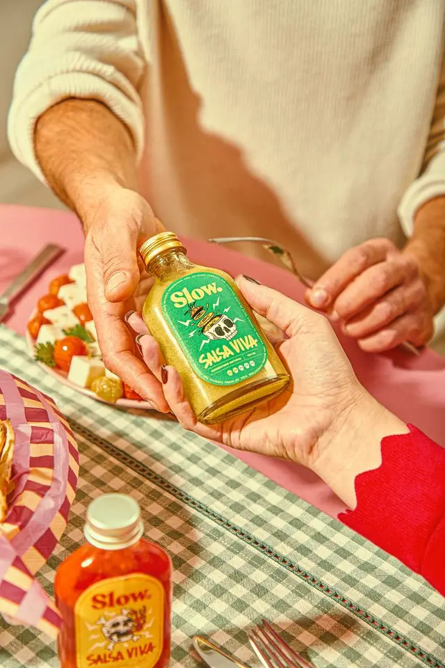 Glass bottle of 'Slow Salsa Viva' passed across pink tabletop with checkered tablecloth