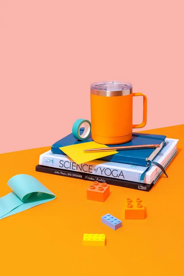 Orange insulated mug on stacked books with colorful desk accessories