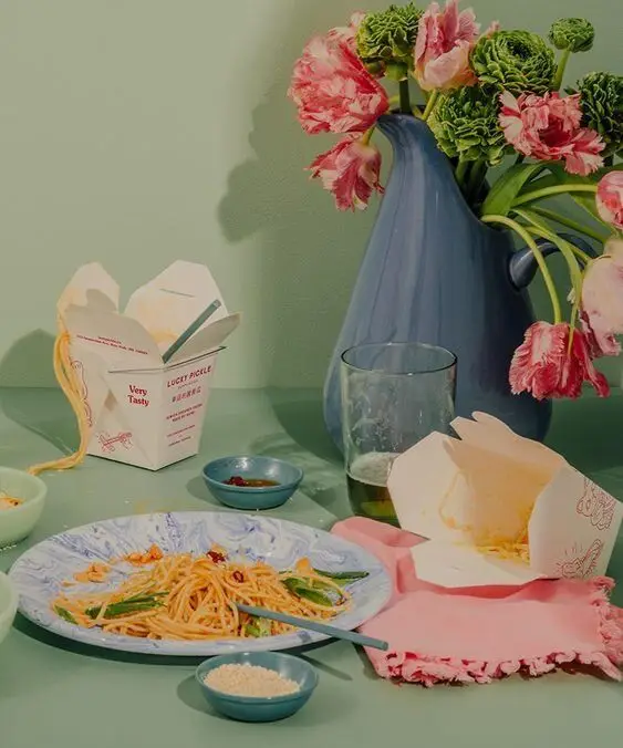 Pastel tabletop still life with spaghetti, takeout boxes and blue pitcher of flowers