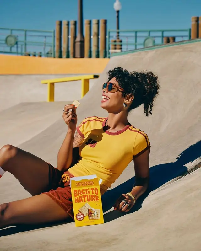 Woman snacking on Back to Nature crackers in skatepark bowl, yellow ringer tee