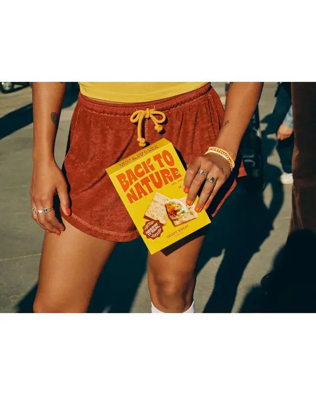 Back To Nature cracker box held with rust terry shorts — sunlit street-style photo