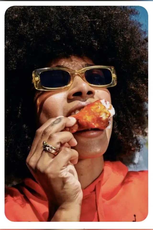 Close-up portrait: gold sunglasses, orange hoodie, biting a saucy chicken wing