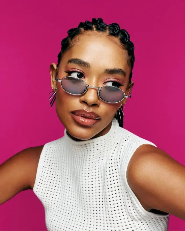 Editorial portrait — cornrow braids, oval sunglasses, white perforated top on magenta back