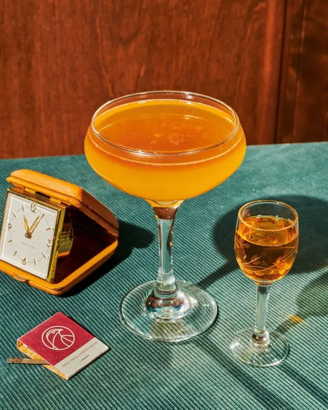 Orange coupe cocktail with cordial and vintage travel alarm clock on teal tablecloth